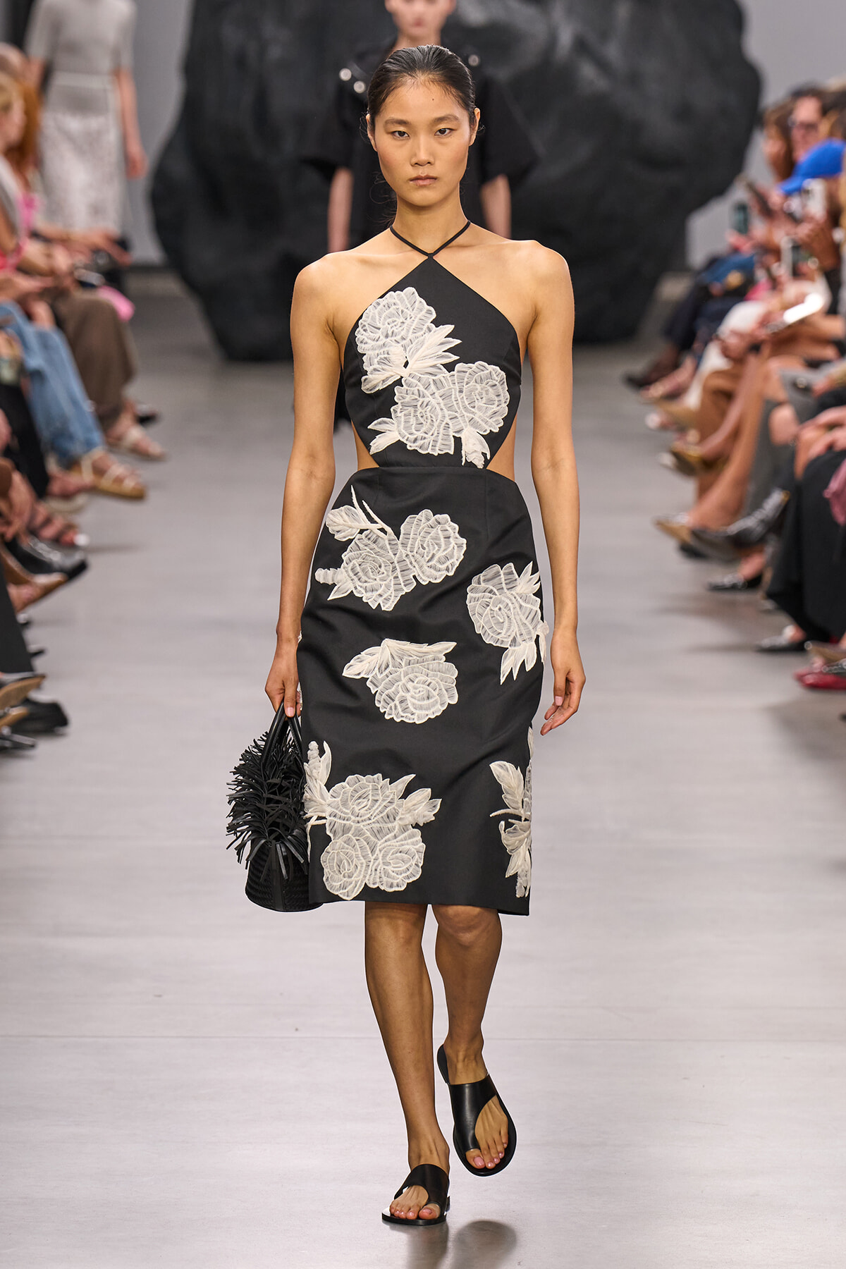 Female model walking a runway in a black halter-neck midi dress with large white floral embroidery, carrying a black spiky-fringed handbag.