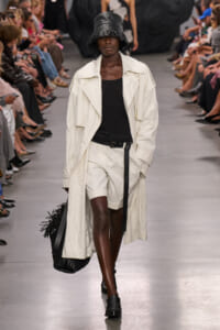 Runway model in a cream trench, black top and white shorts, with a black textured hat and a black fringe tote bag.