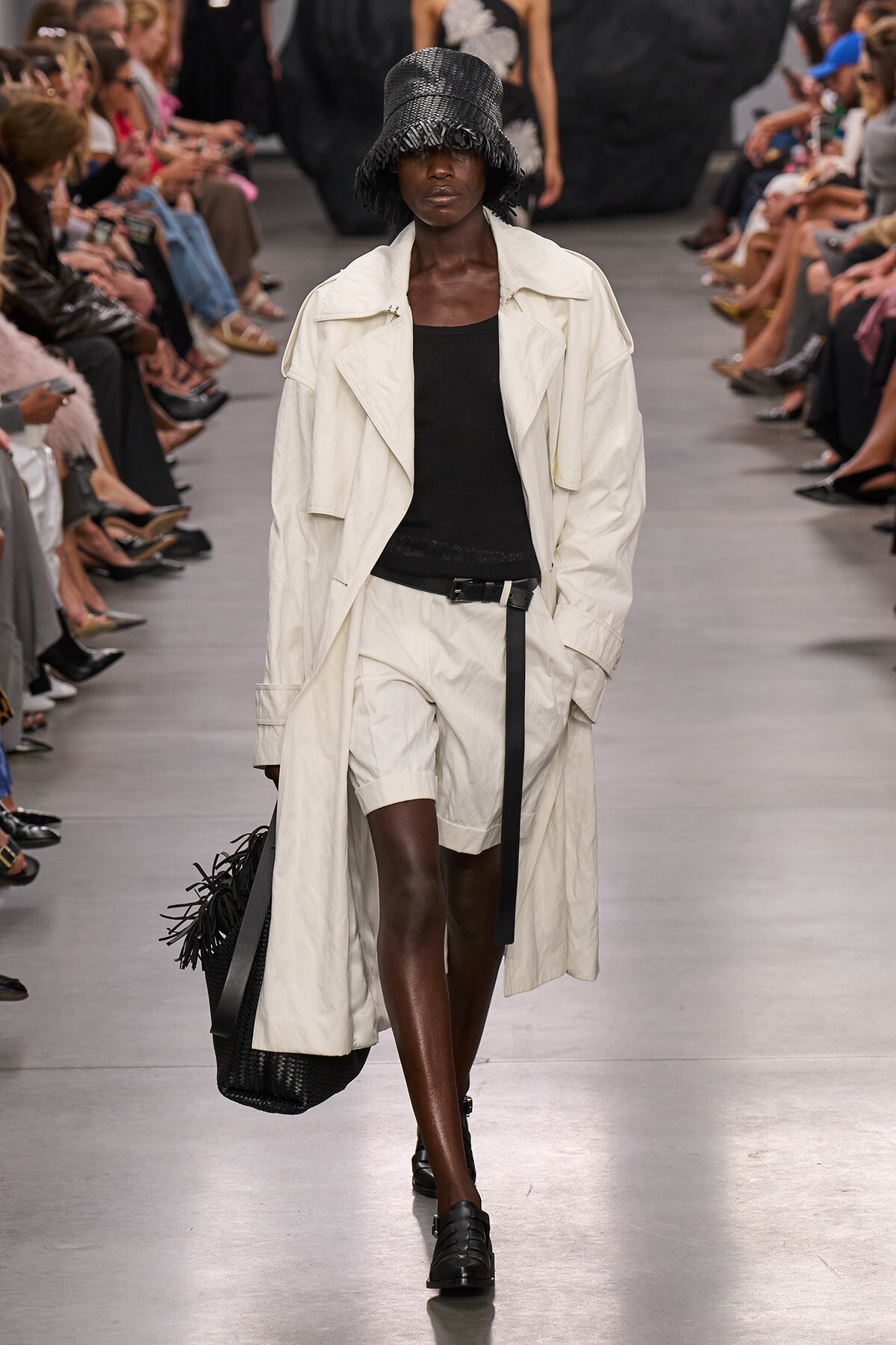 Runway model in a cream trench, black top and white shorts, with a black textured hat and a black fringe tote bag.