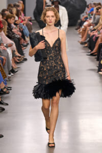 Model on a fashion runway in a black lace slip dress with delicate straps and a feathered fringe hem, clutch in hand.