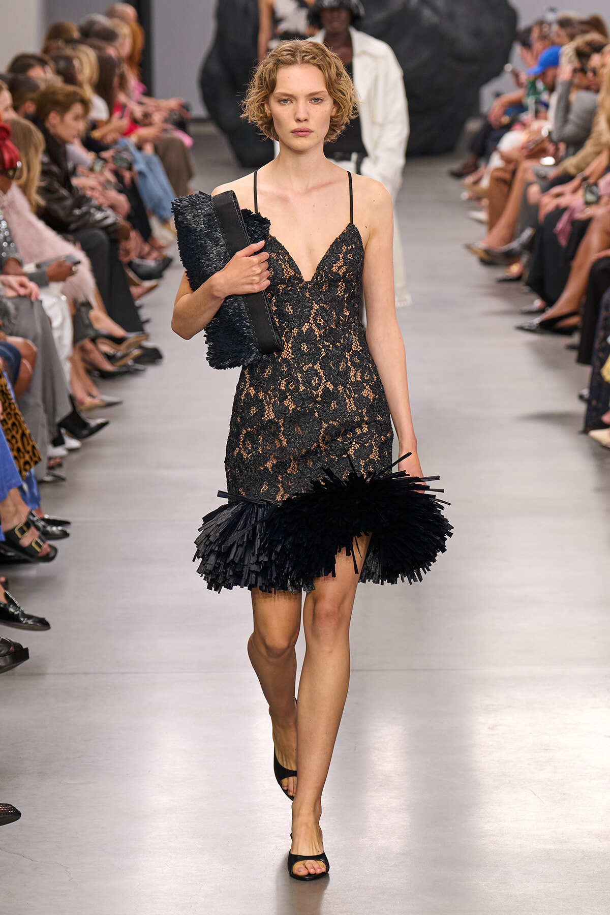 Model on a fashion runway in a black lace slip dress with delicate straps and a feathered fringe hem, clutch in hand.