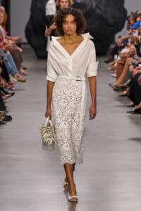 Model walks the runway in a white button-down blouse and ivory lace midi skirt with a belt, carrying a fringed handbag.