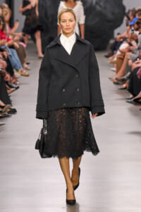 Runway look: model wearing a black double-breasted coat over a white shirt, black lace midi skirt, black heels, and a black handbag on her arm.