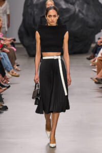 Runway model wearing a black sleeveless knit crop top with a high-waisted pleated black midi skirt and white belt, carrying a small black handbag and wearing white heels.