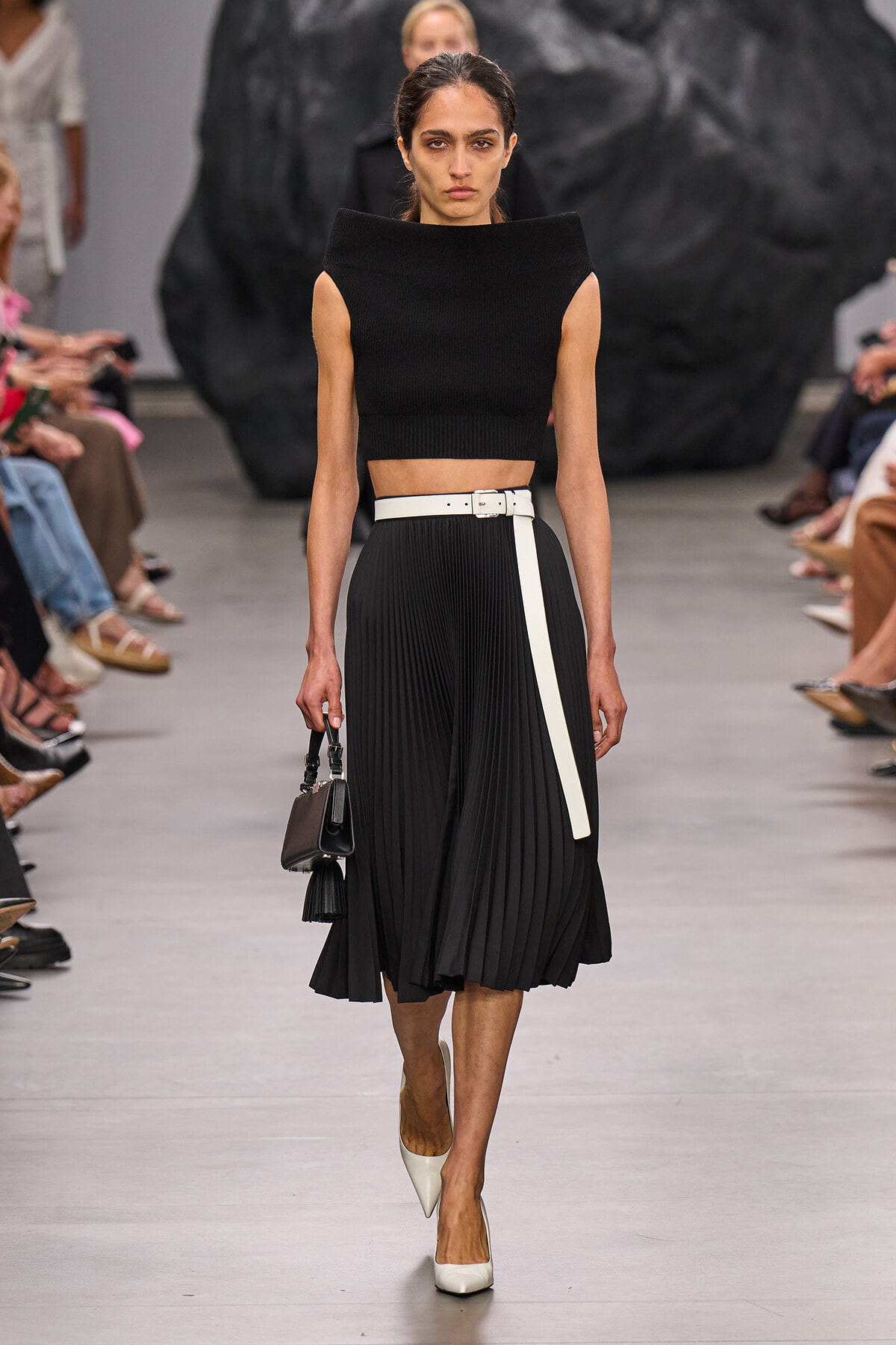 Runway model wearing a black sleeveless knit crop top with a high-waisted pleated black midi skirt and white belt, carrying a small black handbag and wearing white heels.