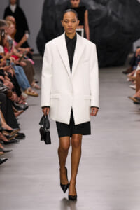 Model walking a runway in an oversized white blazer over a black shirt and skirt, carrying a small black handbag.