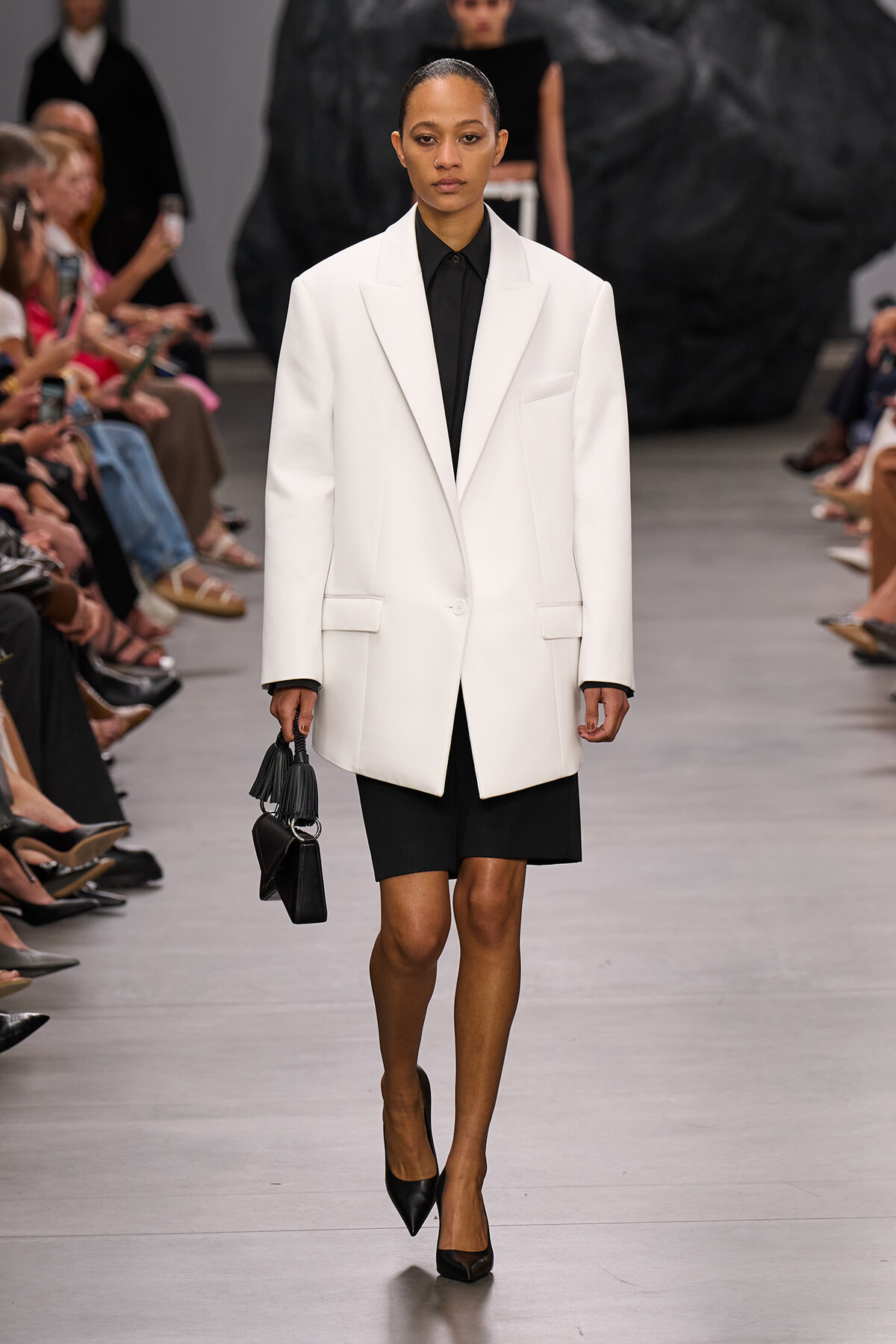 Model walking a runway in an oversized white blazer over a black shirt and skirt, carrying a small black handbag.