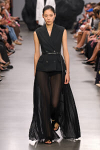 Model on a runway in a black sleeveless double-breasted blazer with a slim belt and sheer pleated black skirt, walking forward.