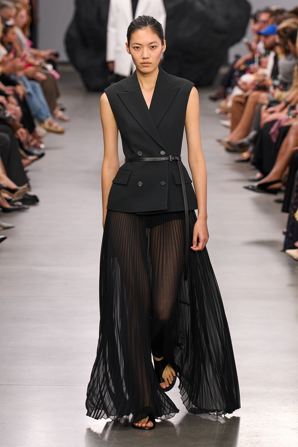 Model on a runway in a black sleeveless double-breasted blazer with a slim belt and sheer pleated black skirt, walking forward.