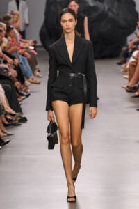 Model walks the runway in a black tailored blazer romper with a belt and black handbag with tassels.