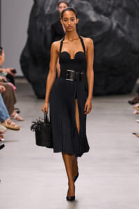 Model walks a runway in a black strapless bustier top, wide pleated midi skirt with a front slit, a chunky belt, and a black woven handbag with fringe.