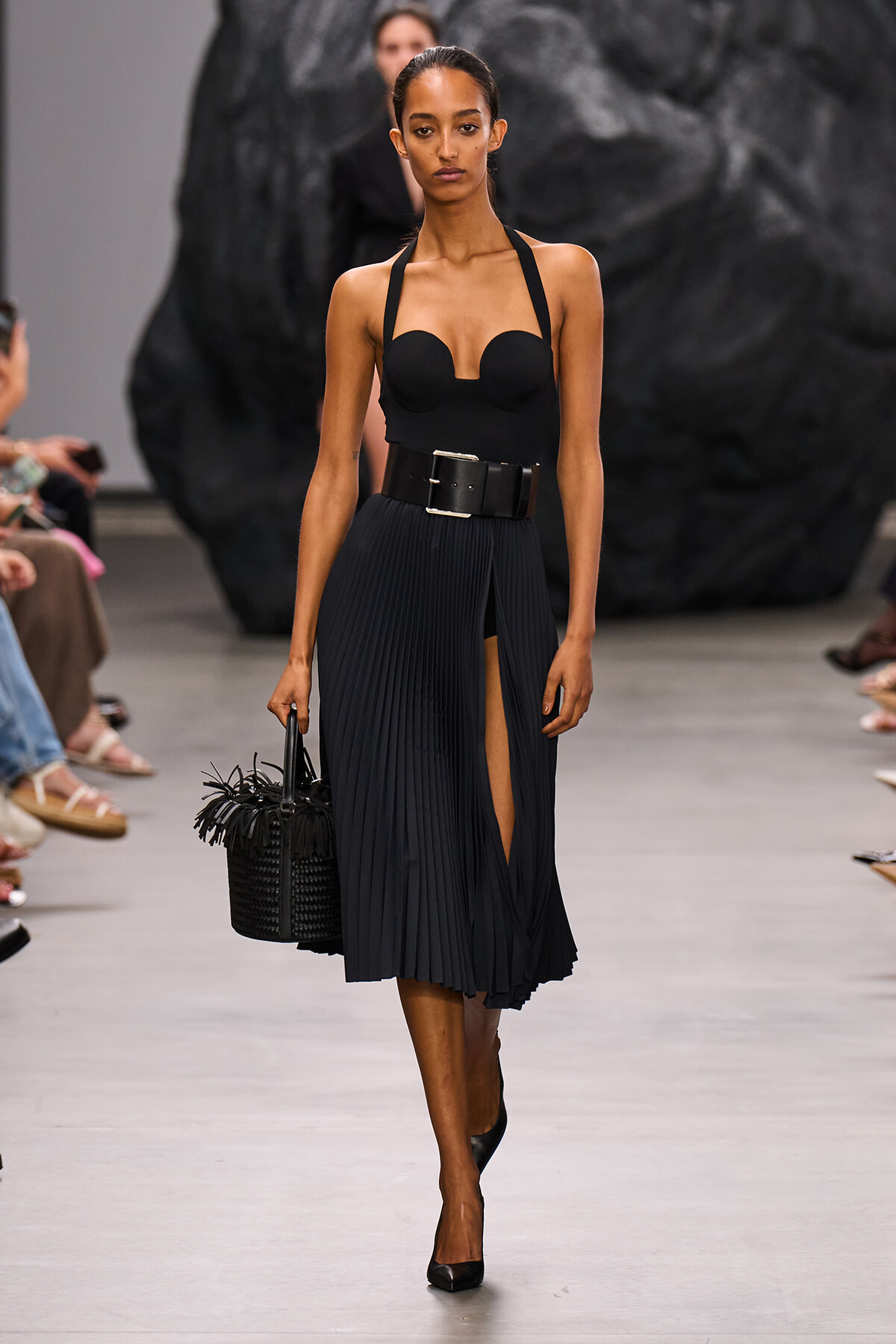 Model walks a runway in a black strapless bustier top, wide pleated midi skirt with a front slit, a chunky belt, and a black woven handbag with fringe.