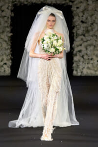 Bride model on a runway in an ivory beaded gown and long veil, holding a white bouquet as she walks.