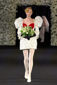 Runway model wearing an ivory bejeweled mini dress with oversized puff sleeves, a red heart-shaped bodice, net veil, and white bouquet on a dark runway backdrop.