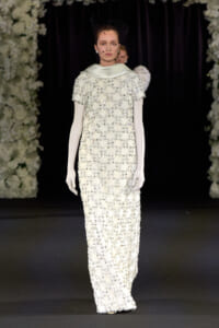 Runway model in an ivory, floor-length beaded gown with short sleeves, white opera gloves, and a black net veil at a fashion show. The dress is heavily embellished with sparkling beads and sequins, set against floral stage decor.
