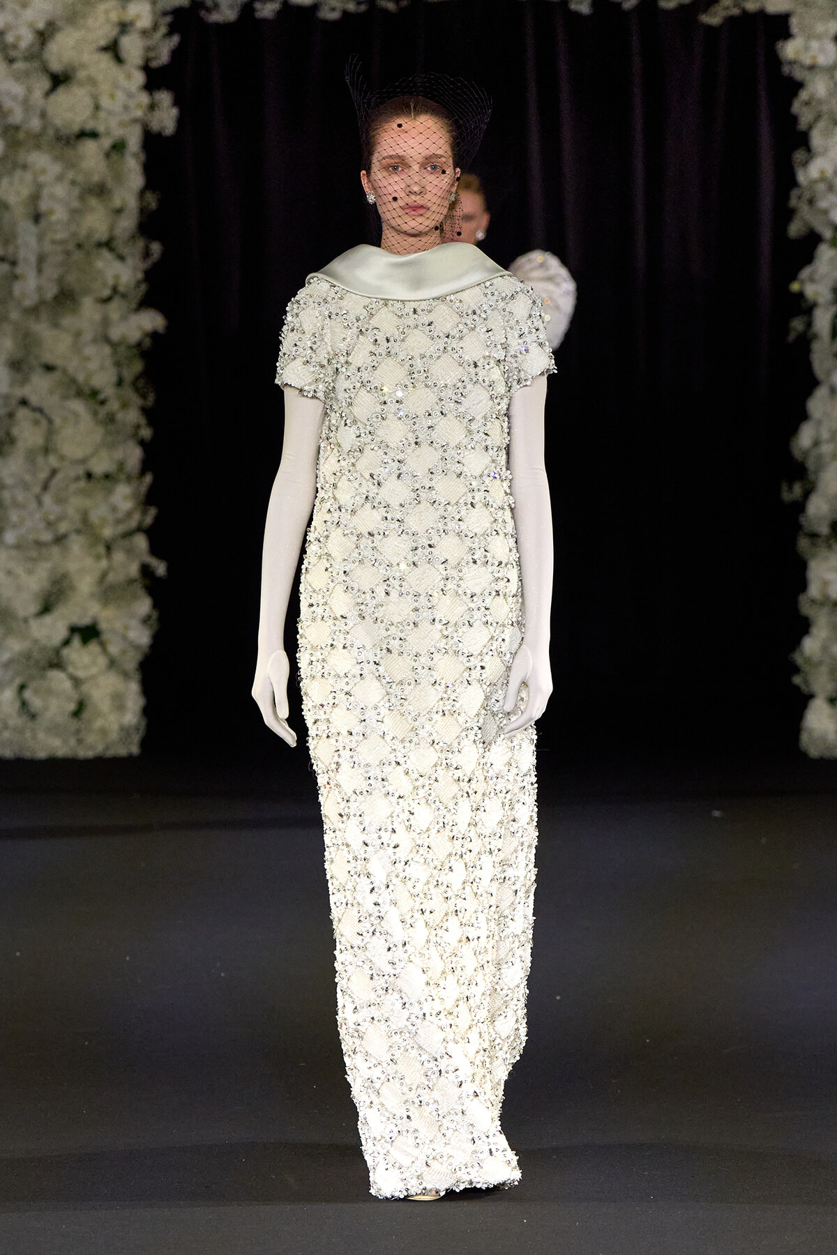 Runway model in an ivory, floor-length beaded gown with short sleeves, white opera gloves, and a black net veil at a fashion show. The dress is heavily embellished with sparkling beads and sequins, set against floral stage decor.
