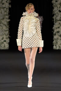 Model walking a runway in an ivory lattice dress with oversized satin ruffles at the neck and large cuffed sleeves, white heels.