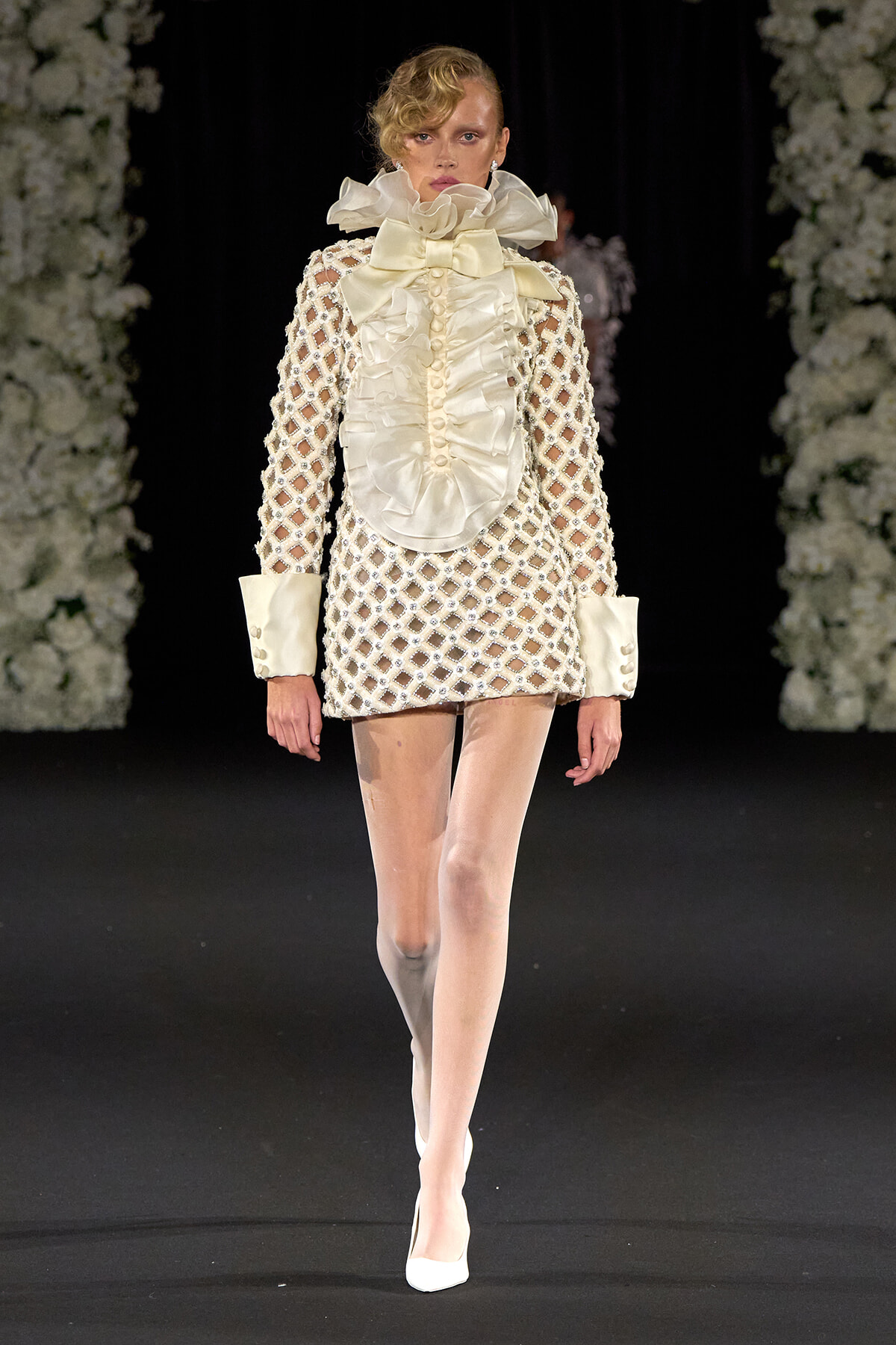 Model walking a runway in an ivory lattice dress with oversized satin ruffles at the neck and large cuffed sleeves, white heels.