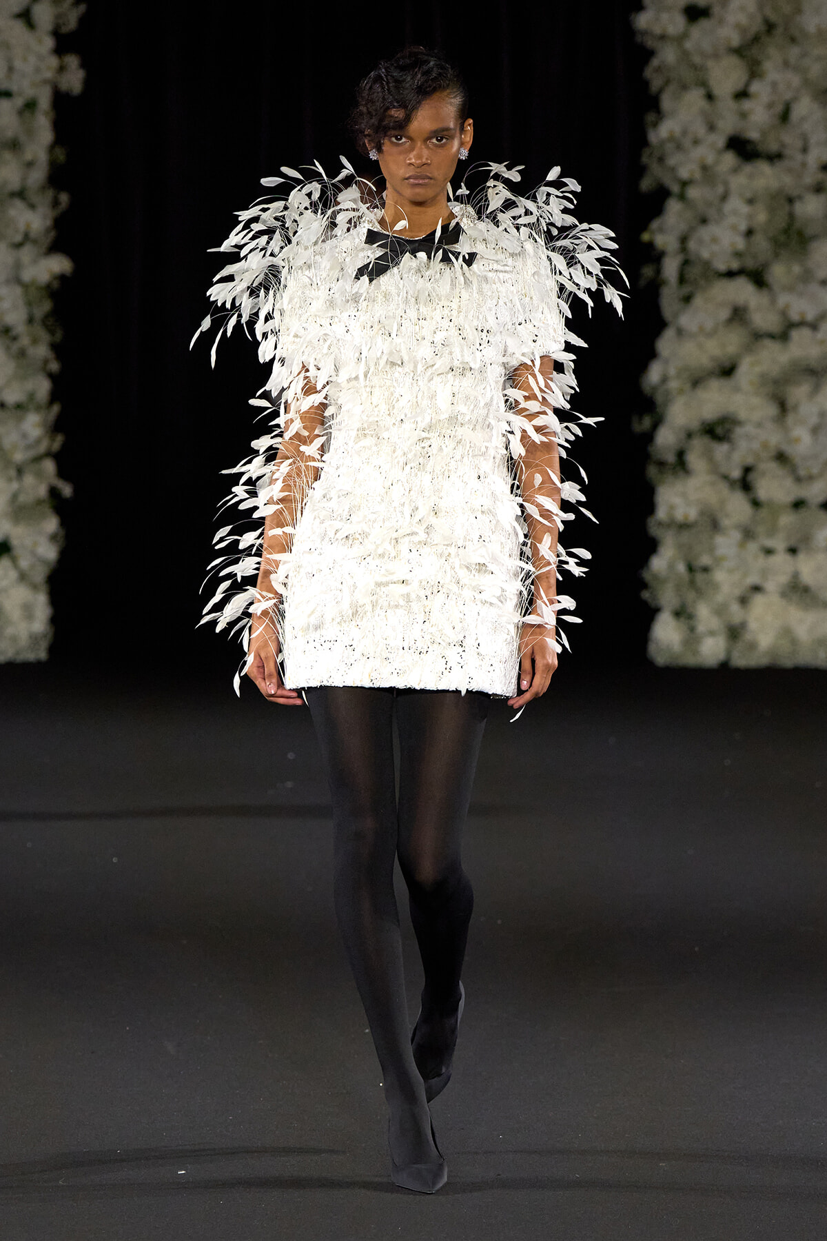 Woman model walks runway in a white feathered, fringed tunic paired with black tights and heels, against a floral backdrop.