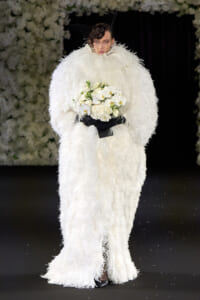 Model walking the runway in an oversized white feathered gown, holding a bouquet of white orchids.