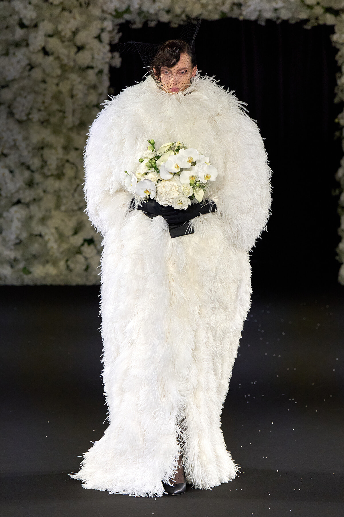 Model walking the runway in an oversized white feathered gown, holding a bouquet of white orchids.