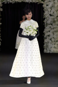 Model walking a runway in a white bead-embellished wedding gown with black gloves, a veil, and a white bouquet.