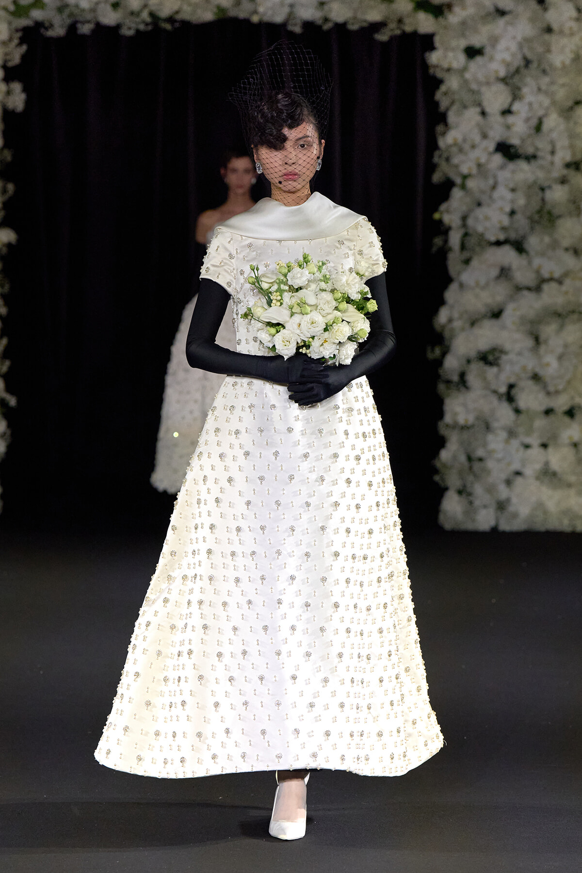 Model walking a runway in a white bead-embellished wedding gown with black gloves, a veil, and a white bouquet.