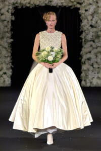 Model on a runway wearing an ivory satin ball gown with a beaded lace bodice and birdcage veil, holding a white bouquet with greenery.