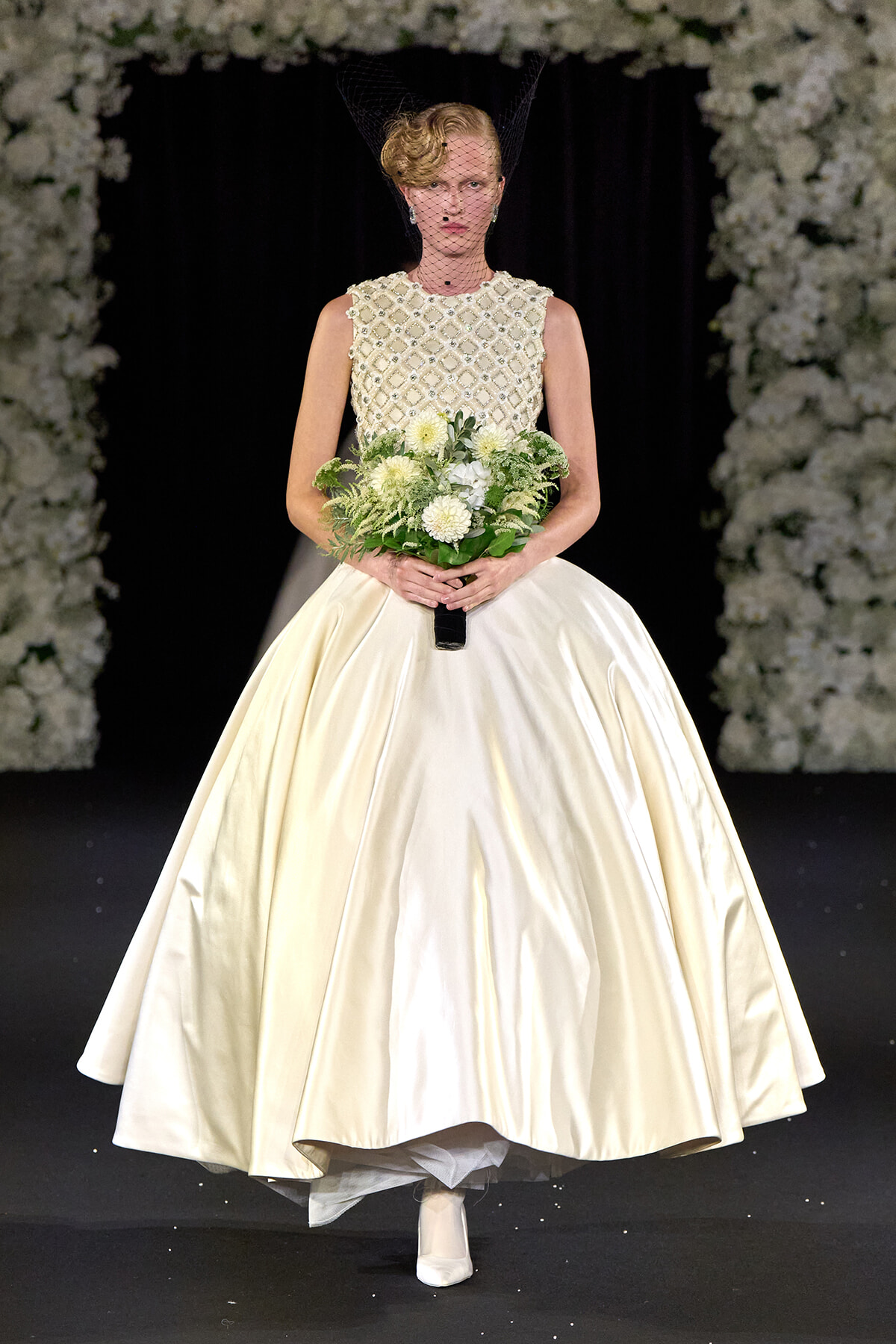 Model on a runway wearing an ivory satin ball gown with a beaded lace bodice and birdcage veil, holding a white bouquet with greenery.