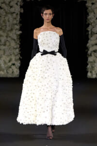 Model on a runway wearing an off-the-shoulder white floral ball gown with a black ribbon belt, black gloves, and a netted veil.
