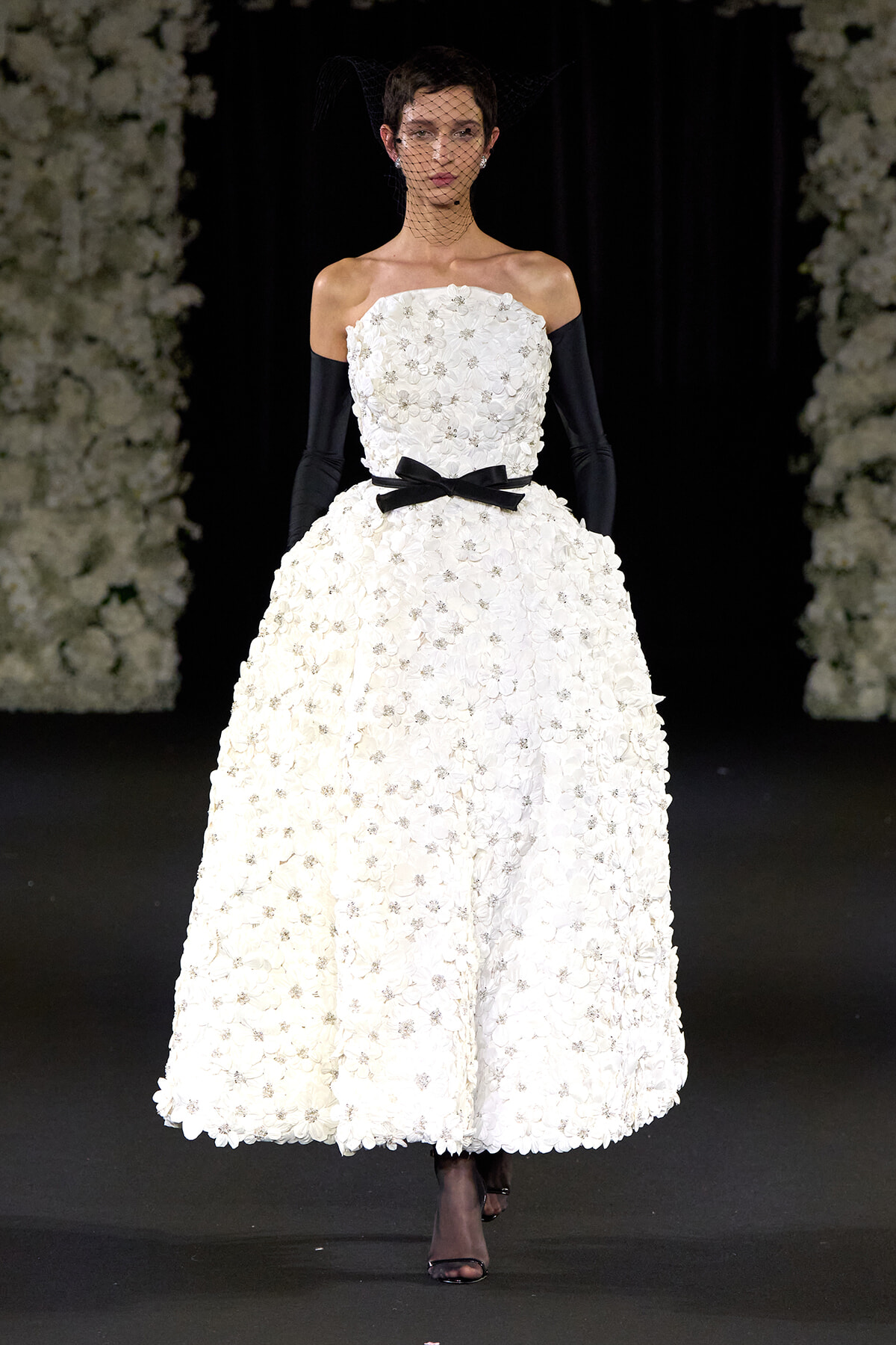 Model on a runway wearing an off-the-shoulder white floral ball gown with a black ribbon belt, black gloves, and a netted veil.