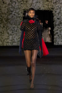 Model walking the runway in a black lattice mini dress with a red rose at the near neckline, sheer veil, and navy gloves with black heels." (informative)