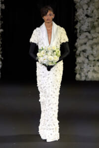 Model on a runway in a white, flower-covered gown with a satin shawl, black gloves, veil, and a white bouquet.