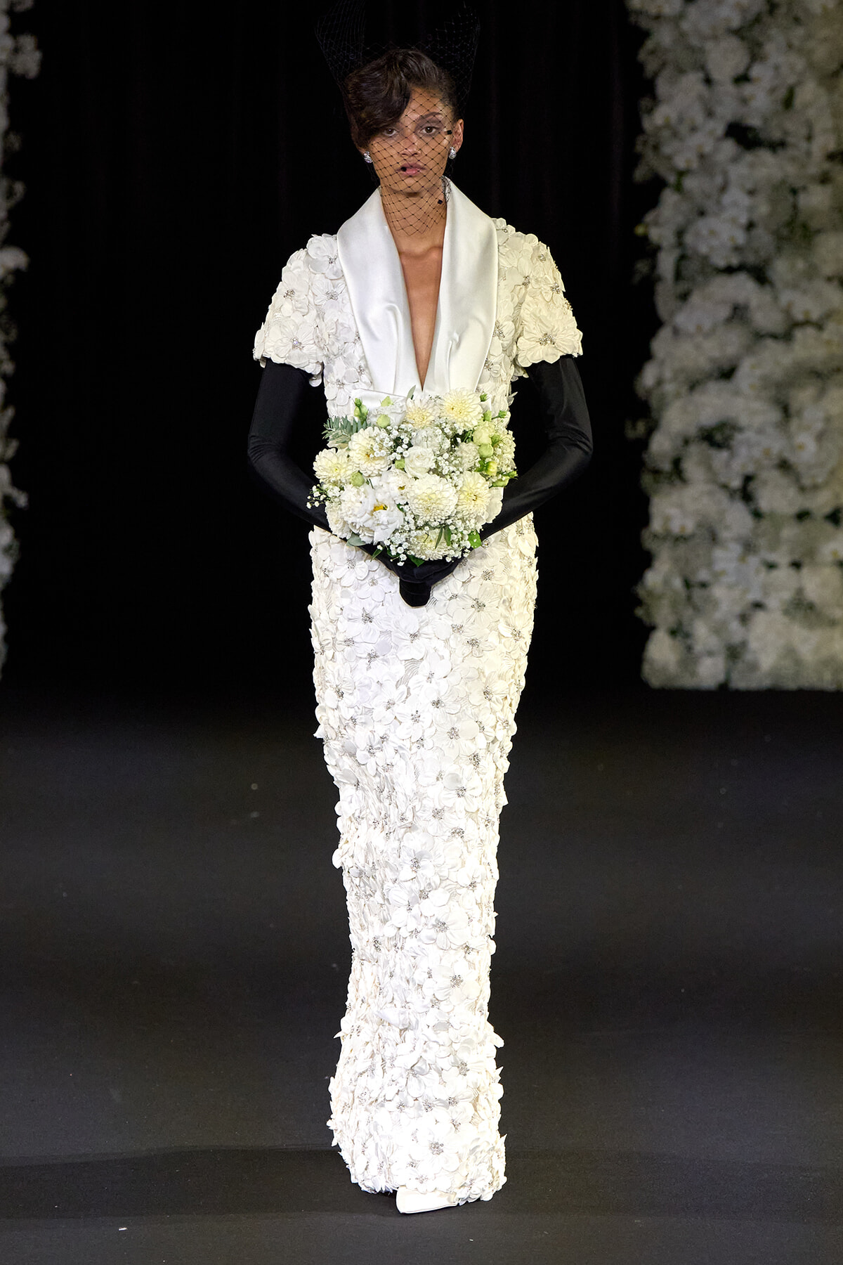 Model on a runway in a white, flower-covered gown with a satin shawl, black gloves, veil, and a white bouquet.