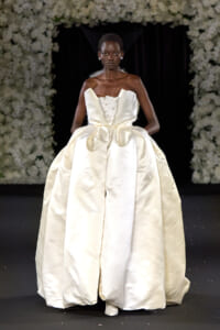 Model walks the runway in an ivory satin ball gown with a sculptural bodice and voluminous skirt, against a floral backdrop.