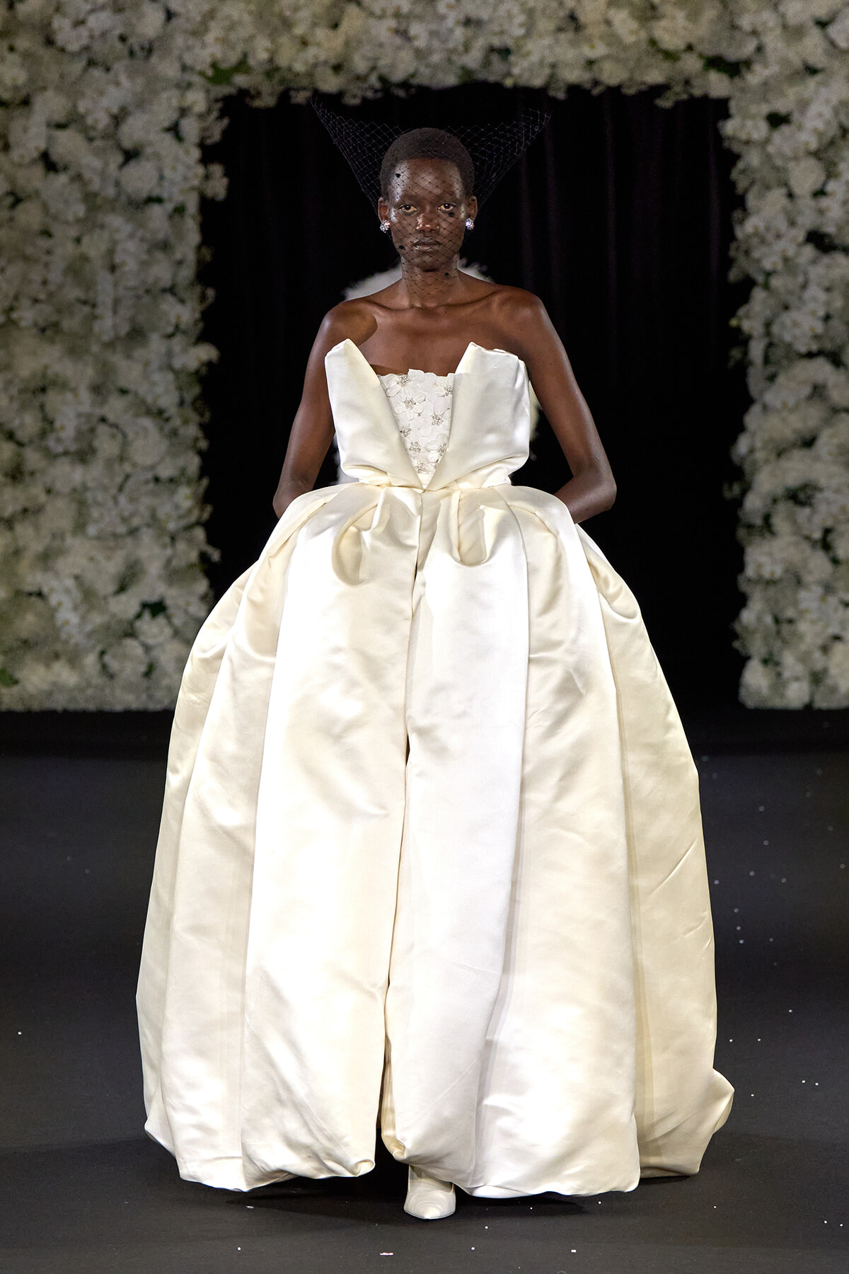 Model walks the runway in an ivory satin ball gown with a sculptural bodice and voluminous skirt, against a floral backdrop.