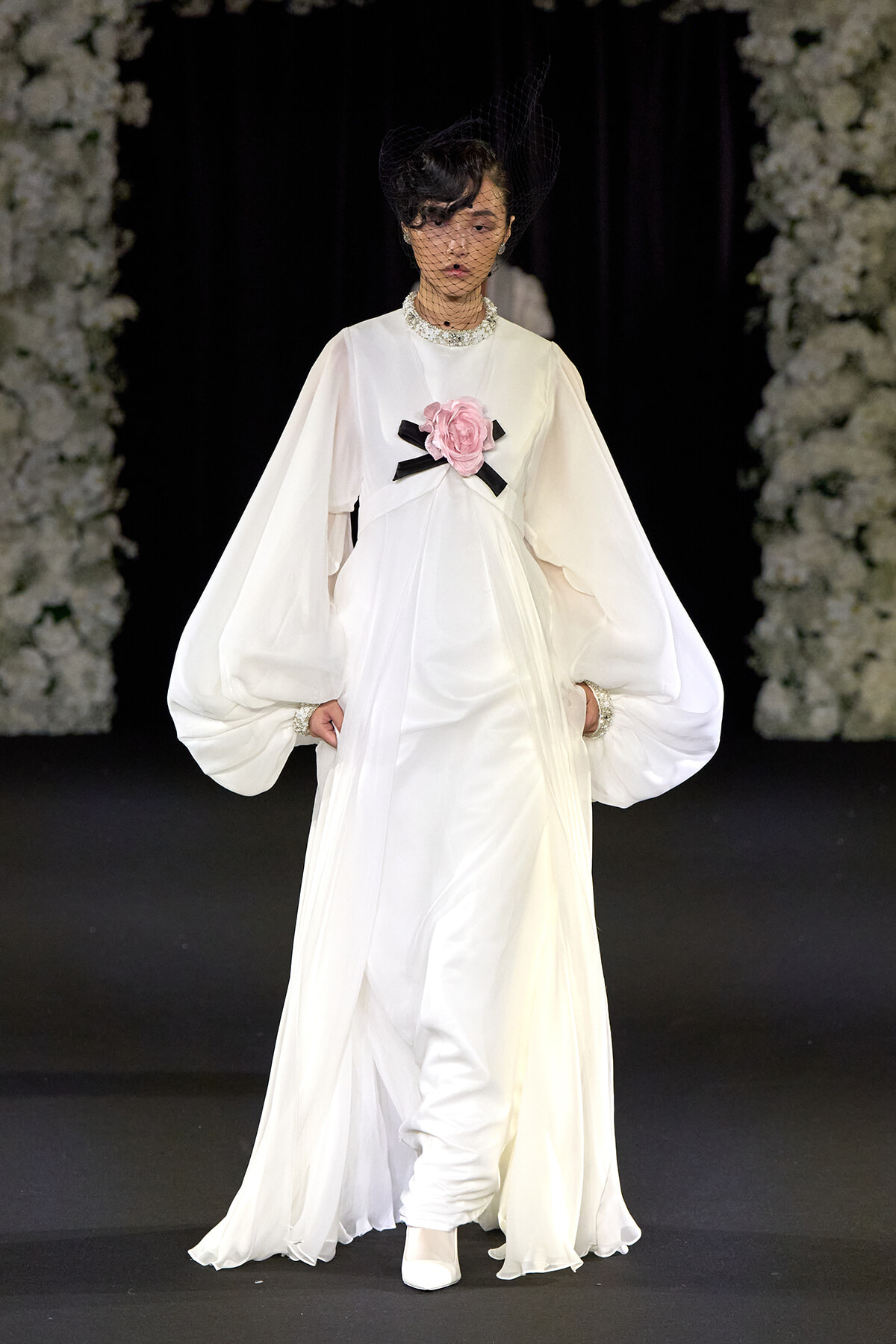 Model on a runway wearing a white, floor-length gown with dramatic balloon sleeves and a pink flower brooch at the chest.
