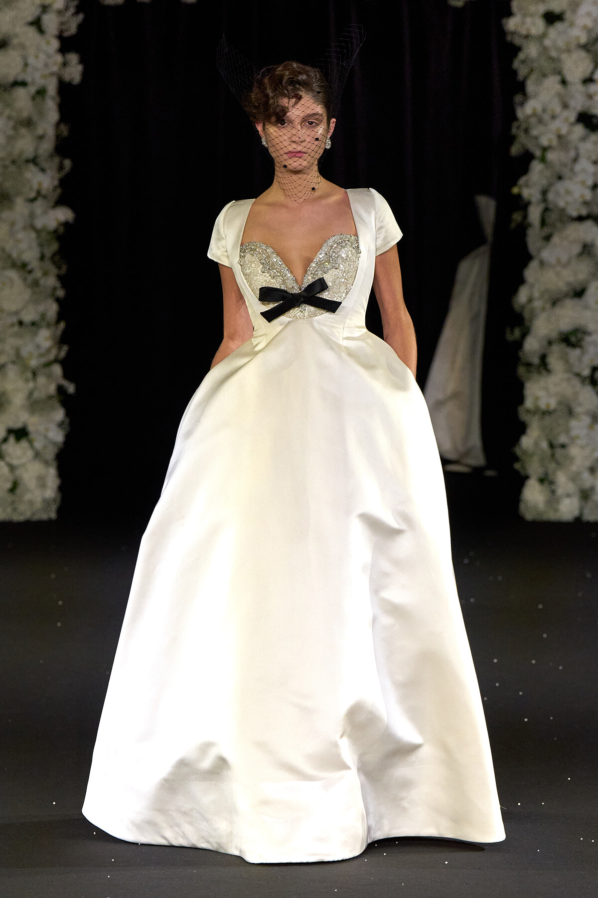 Model on runway wearing a white satin ballgown with a beaded bodice, short sleeves, and a black bow at the waist, plus a decorative veil headpiece.