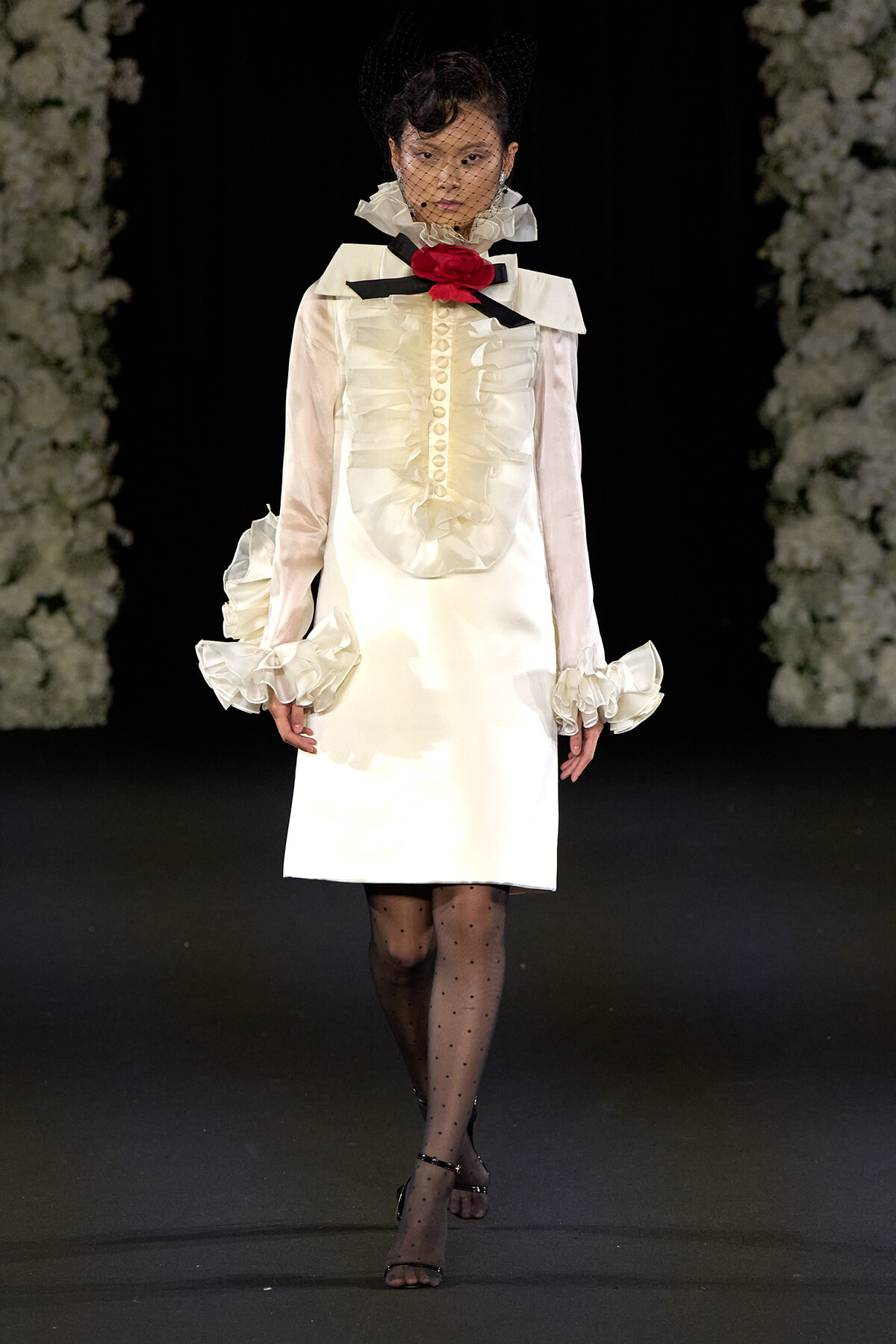 Runway model in a cream satin dress with ruffled front, oversized collar, black bow and red rose, and a black net veil.