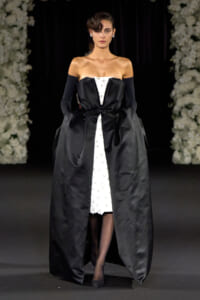 Model walking a runway in an off-shoulder black satin gown with a white lace center panel, oversized bow at the waist, and a voluminous outer skirt; she wears long black gloves and small earrings.
