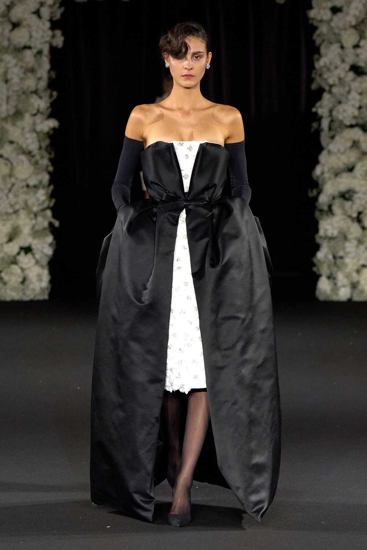 Model walking a runway in an off-shoulder black satin gown with a white lace center panel, oversized bow at the waist, and a voluminous outer skirt; she wears long black gloves and small earrings.