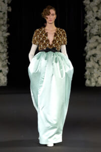 Model on runway wearing a brown beaded cape-like top with a deep V and a mint satin full skirt cinched by a black bow belt.