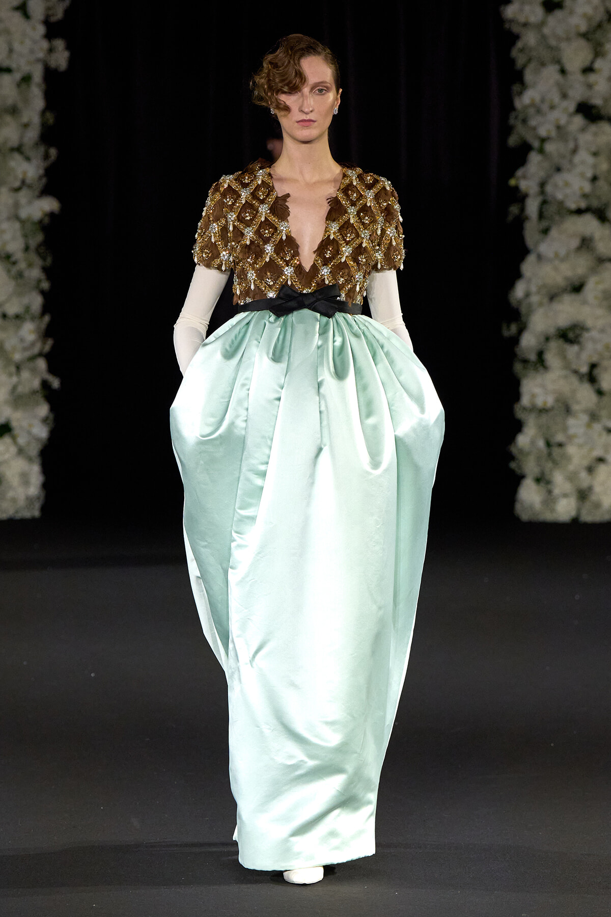 Model on runway wearing a brown beaded cape-like top with a deep V and a mint satin full skirt cinched by a black bow belt.