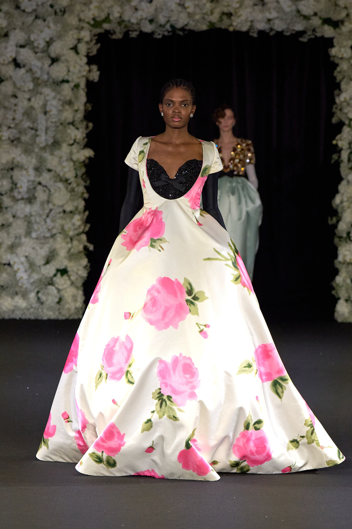 Model walks the runway in a cream ballgown with large pink rose prints and a black sequined sweetheart bodice.