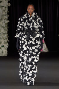 Runway model in a black-and-white floral appliqué suit with a large black bow at the waist, wearing long black gloves.