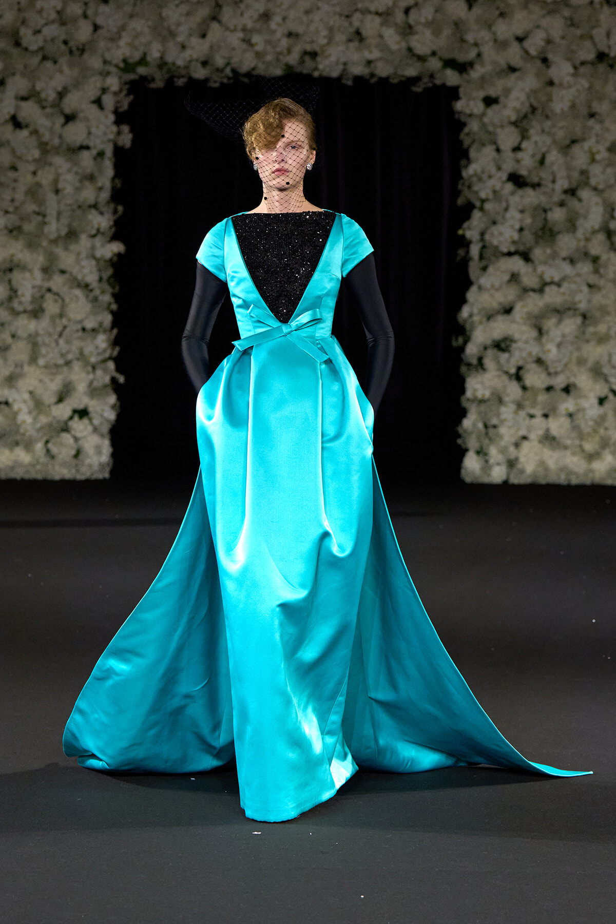 Model walks runway in a turquoise satin gown with black sequined bodice and long black gloves, veil overhead.