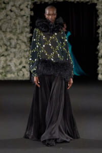Model walking a runway in a black sheer floral blouse with a large ruffle collar and feathered waist, paired with a long black skirt.