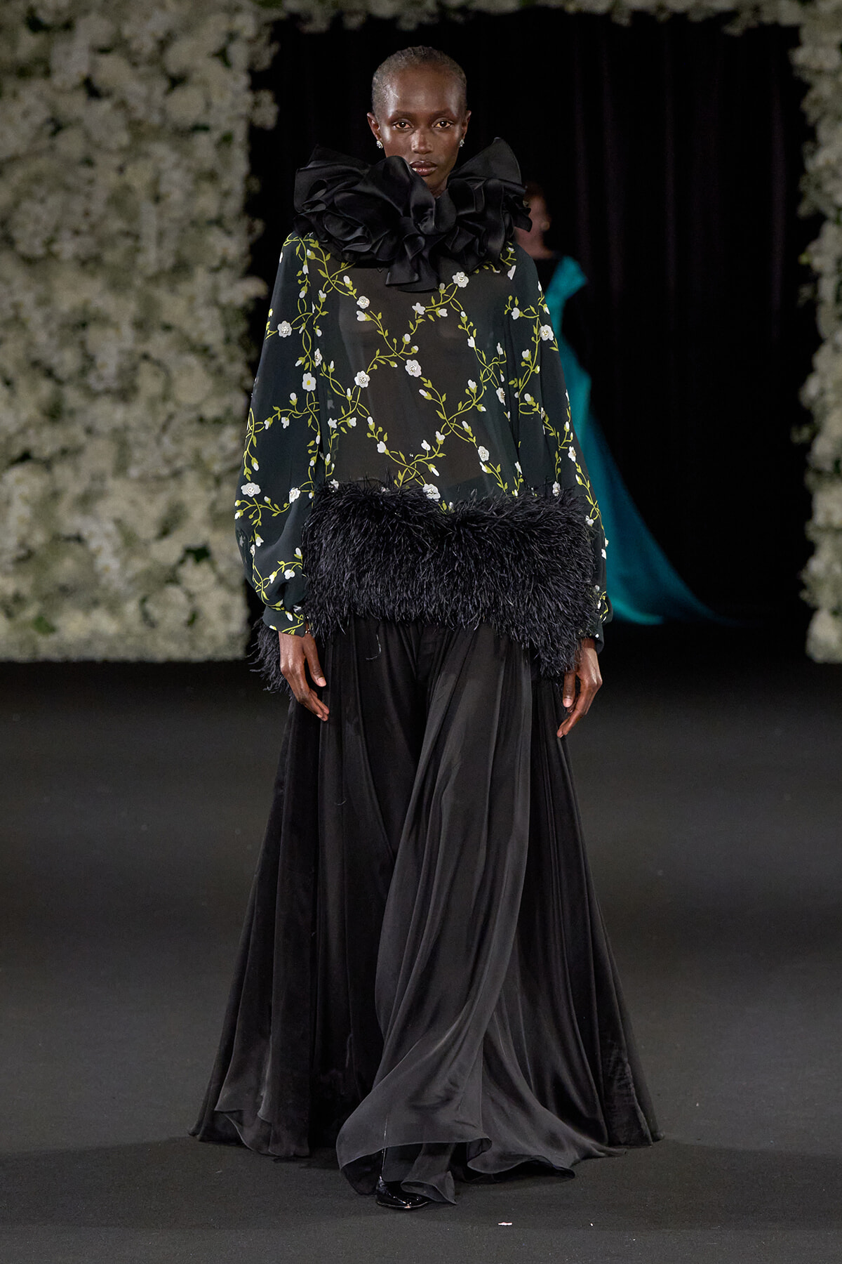 Model walking a runway in a black sheer floral blouse with a large ruffle collar and feathered waist, paired with a long black skirt.
