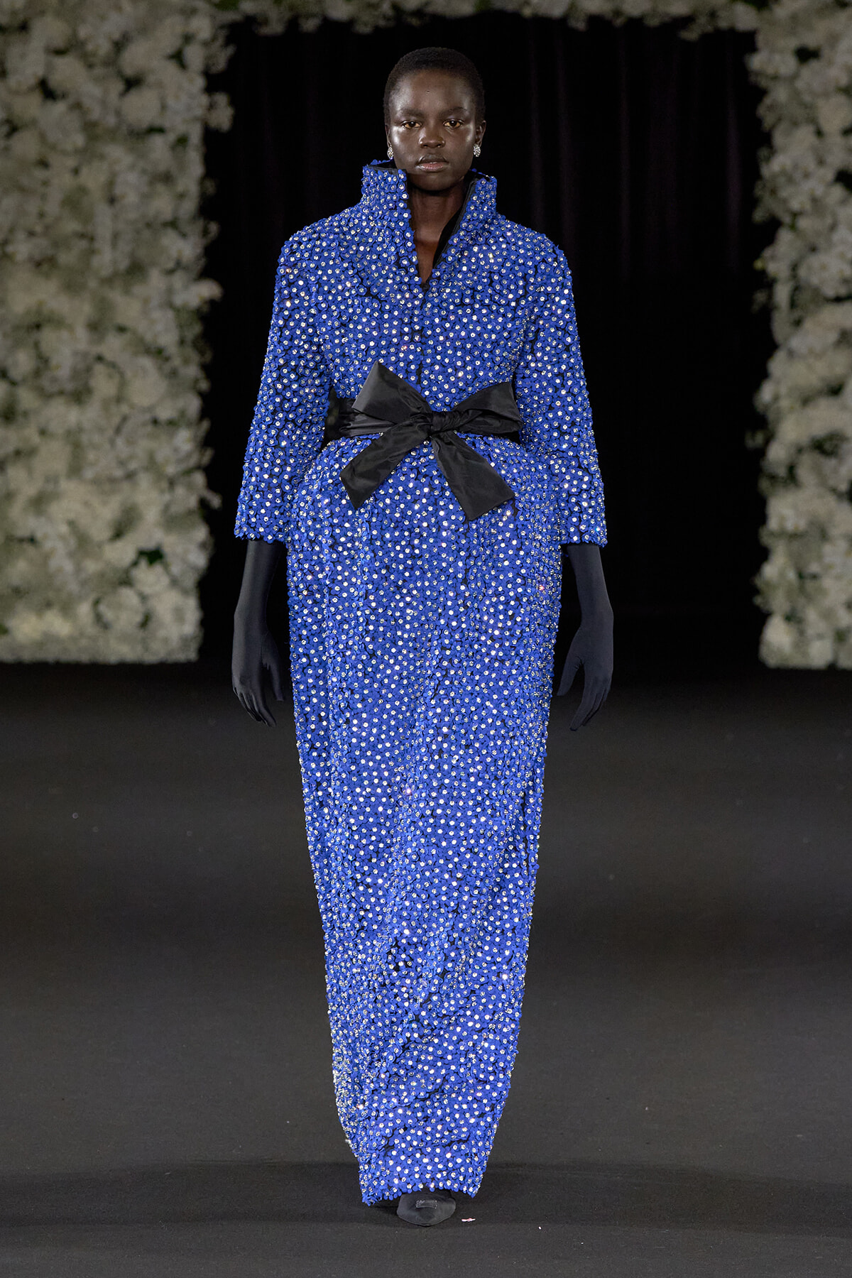 Model walks a runway wearing a floor-length blue beaded dress with a large black bow belt, paired with black gloves against a dark backdrop with floral arrangements.