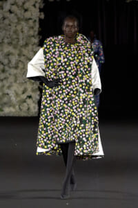 Model on a runway wearing a floor-length black floral dress with pink, yellow, and white flowers, white voluminous sleeves, black gloves, and a black net headpiece.